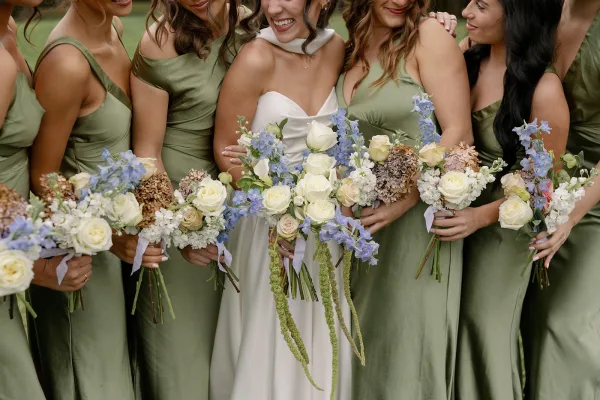 Bridesmaid group photo with bride in strapless gown, bridesmaids in sage green dresses holding white rose and blue delphinium bouquets on a lawn