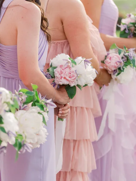 Bridesmaid bouquets of pink and white peonies with greenery, wrapped in white ribbon, held by lavender and blush dressed bridesmaids outdoors