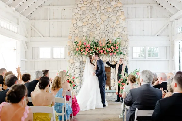 Bride and groom share their wedding first kiss before a fireplace ceremony backdrop, her veil catching the light against white barn walls.