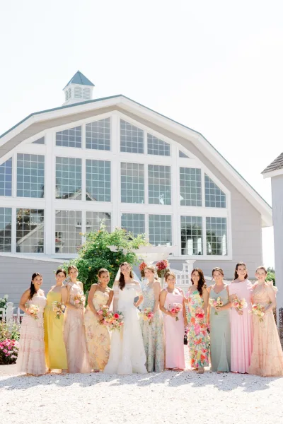 Bridal party portrait of the bride and bridesmaids holding colorful bouquets, posed by a shingle building with large windows and a garden pergola.
