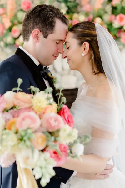 Couple portrait in a forehead touch pose, bride holding a colorful bouquet as the groom embraces, framed by a floral backdrop.