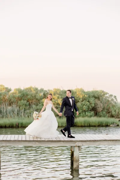 Couple portrait of bride and groom walking hand in hand on a wooden dock by a calm lake, her pastel bridal bouquet catching soft light.
