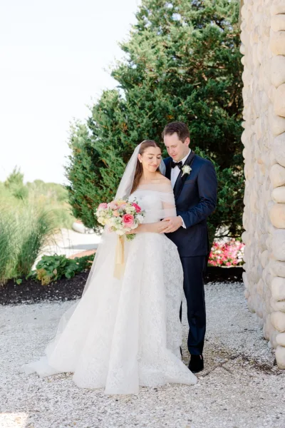 Couple portrait with the bride holding a bouquet and a long veil, the pair standing by a stone wall beside an evergreen tree and gravel path