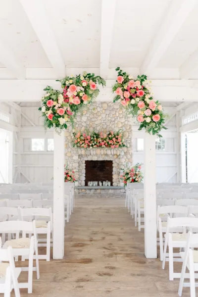 Ceremony setup centers on a fireplace wedding altar against a stone wall, with candles in a white barn interior with wooden floors.