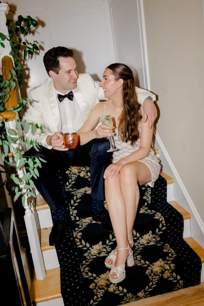 Couple portrait of a bride and groom with drinks, laughing on a staircase as he wraps an arm around her, a copper mug catching the light.