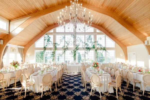 Reception decor with long banquet tables and blush and ivory centerpieces in a hall framed by arched windows and a vaulted wood ceiling.