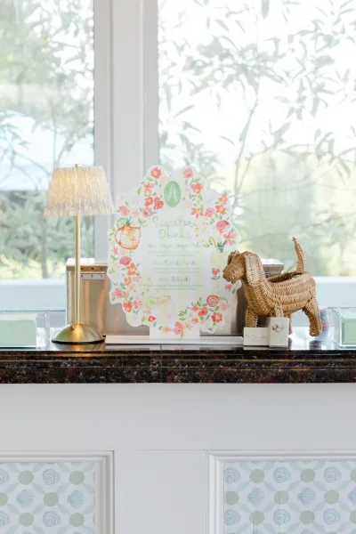 Signature drink sign for the wedding bar menu, beside a wicker dog on a marble countertop by garden-view windows.