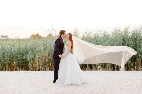 Wedding kiss portrait of a couple holding hands, her long veil trailing as they embrace amid tall grass and reeds under a glowing sunset sky.
