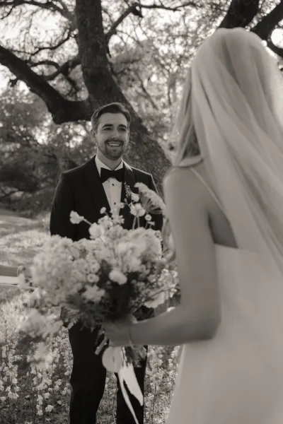 First look moment as bride walks to groom under oak trees on a green lawn, her veil trailing and bouquet in hand, groom in tuxedo smiling