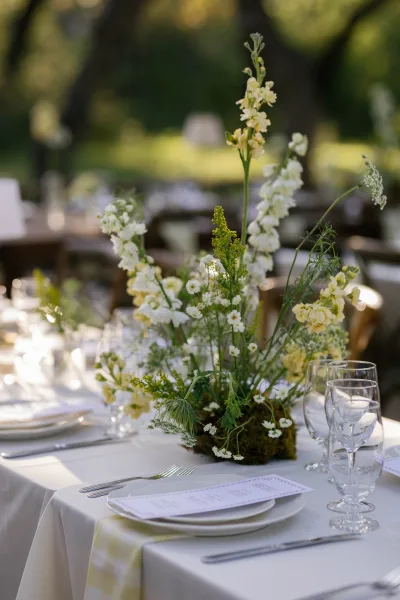 Reception tablescape with wildflower wedding centerpiece on a white tablecloth, plates and glassware set in a sunlit garden under trees