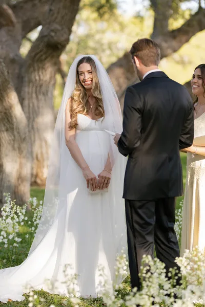 Wedding vows as bride in a long veil reads from a vow booklet to groom in a black suit during an outdoor garden ceremony under trees