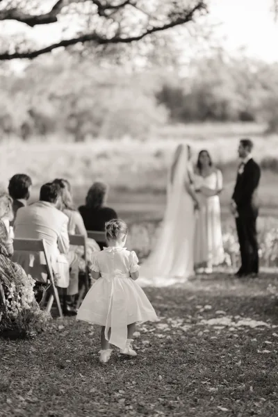 Ceremony moment at an outdoor wedding ceremony as bride in veil and groom in suit stand ahead, flower girl with bow walks petal aisle under tree shade