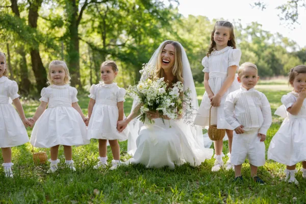 Bridal party portrait of bride with flower girls laughing beside a ring bearer in a sunlit park, holding a wildflower bouquet and veil