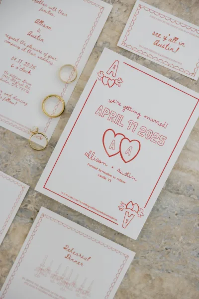 Wedding invitation suite with red wedding invitations and gold rings, featuring scalloped border, monogram, and heart on stone surface