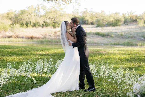 Wedding kiss portrait of bride and groom kissing with a flowing veil and long train in a wildflower meadow by a pond