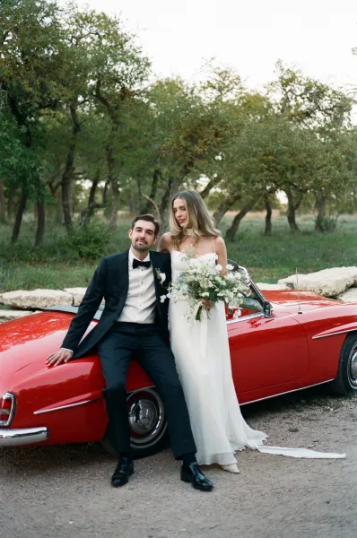 Couple portrait of bride in a strapless wedding dress with bouquet leaning on a vintage convertible as groom in tuxedo sits nearby outdoors