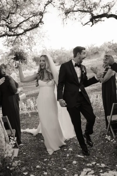 Wedding recessional with bride and groom walking aisle, holding hands as she raises a bouquet; flower petals scatter in an outdoor field