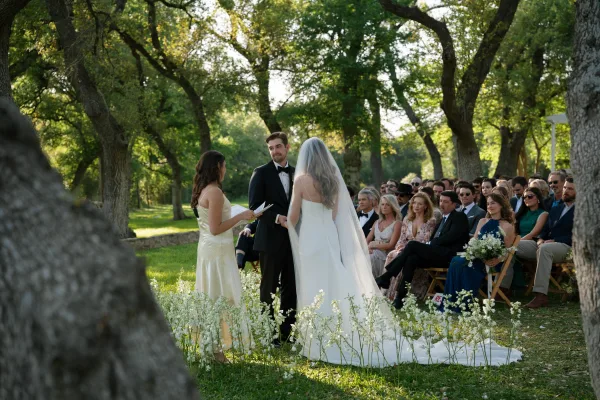 Wedding vows as bride in veil and groom in black tuxedo hold hands while officiant reads from a vow book on a sunlit oak lawn