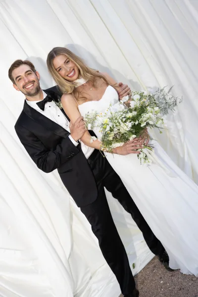 Couple portrait of bride and groom smiling, groom hugging her as she holds a bouquet before a white draped fabric backdrop