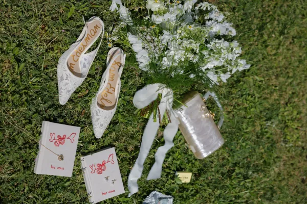 Wedding details flatlay with lace bridal shoes, vow books, white wildflower bouquet, pearl earrings and champagne bottle on grass lawn