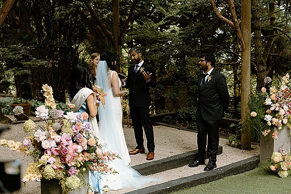 Wedding vows as bride and groom face each other under a wooden pergola, bride in veil holding bouquet, garden trees behind