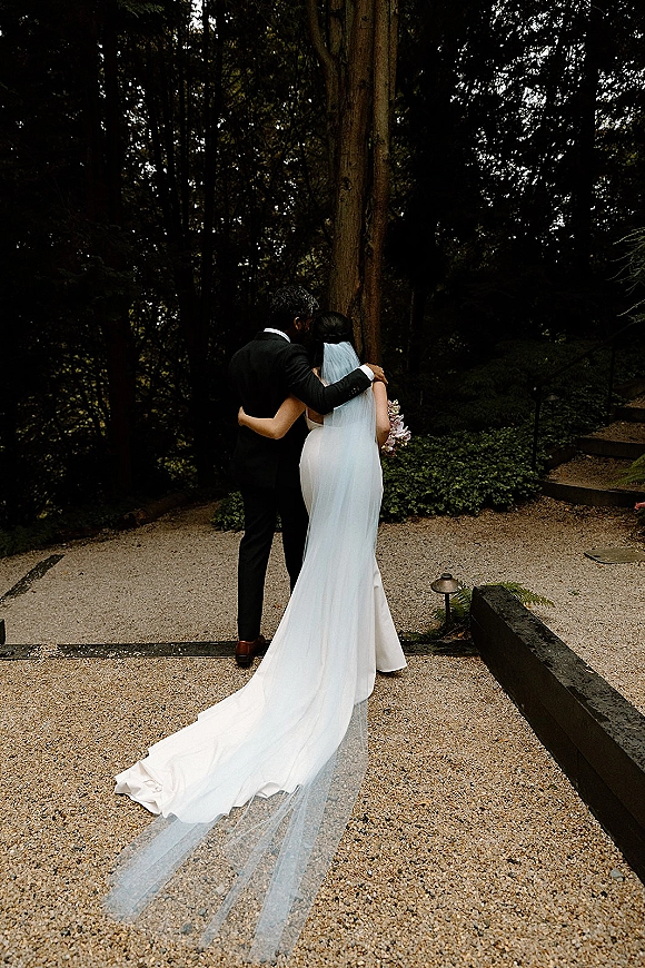Couple portrait of bride and groom from behind walking a gravel forest path, her long veil and train trailing as she holds a bouquet