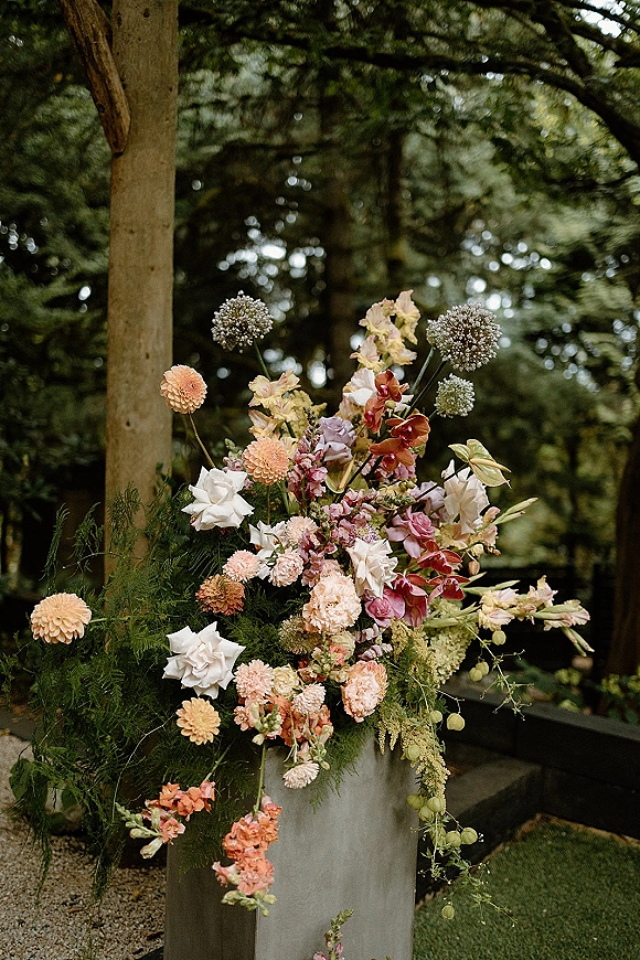 Wedding floral arrangement with white roses and peach dahlias on a metal pedestal stand, set on an outdoor deck by forest trees