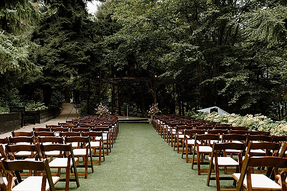 Ceremony setup for an outdoor wedding ceremony with wood folding chairs and green aisle runner leading to a floral wooden arbor in the forest