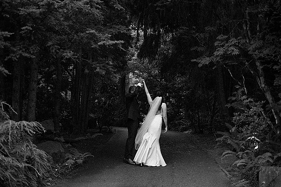 Couple portrait of bride and groom dancing on a forest garden path, her long veil trailing behind his dark suit