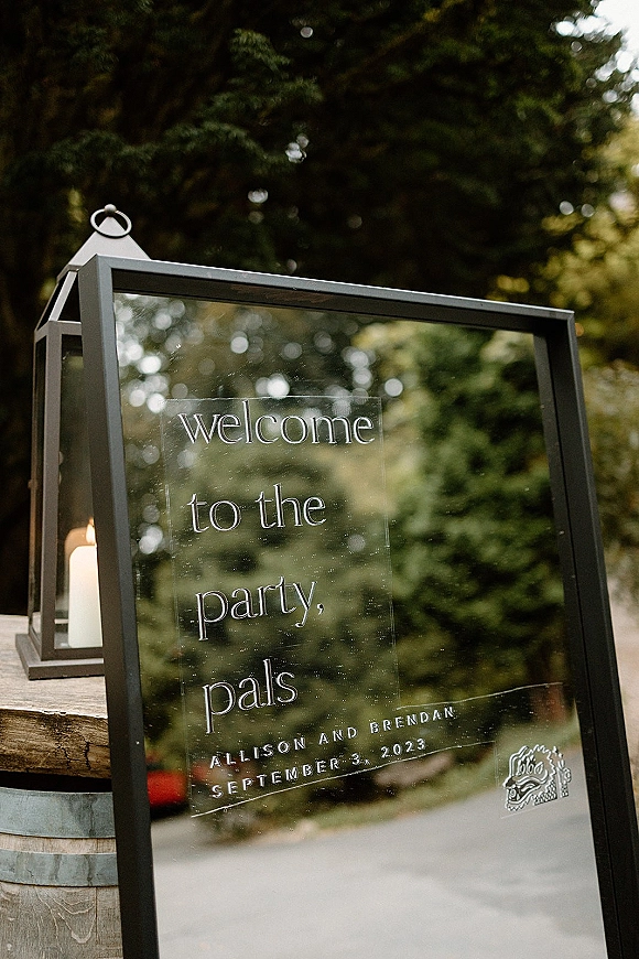 Wedding welcome sign on a wooden barrel with a clear acrylic wedding sign and black lantern with pillar candle along a tree-lined path
