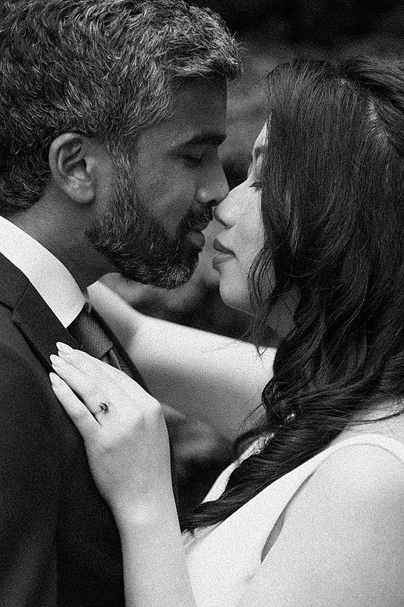 Wedding kiss portrait in black and white of bride holding groom’s face, engagement ring visible as they kiss against a blurred outdoor backdrop