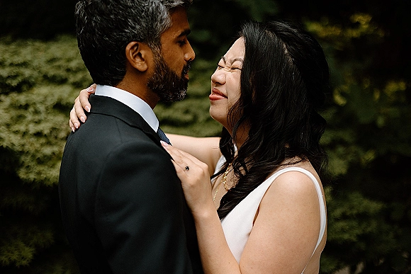 Couple portrait of bride and groom close up, embracing cheek to cheek in greenery, her white dress and his black suit visible
