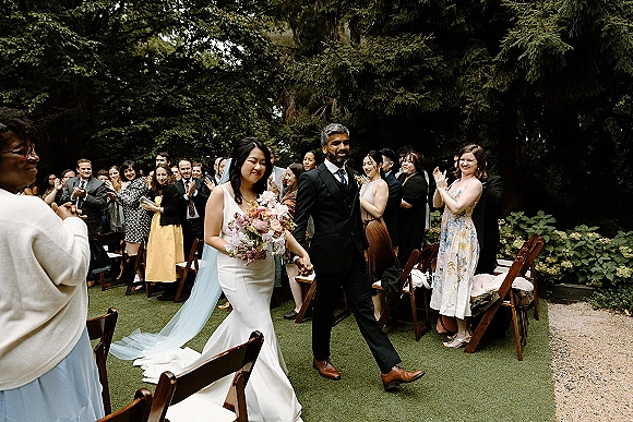Wedding recessional as bride and groom walk the aisle holding hands, guests clapping beside wooden chairs in an outdoor garden setting