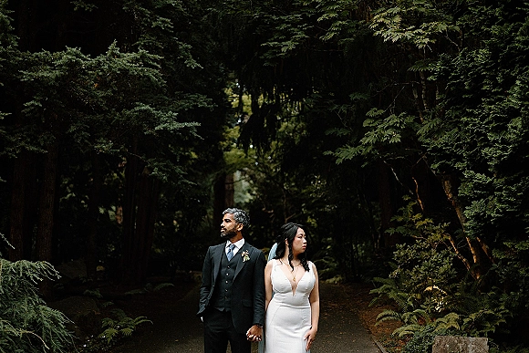 Couple portrait of bride and groom holding hands on a forest path, her veil trailing beside his suit with boutonniere amid ferns