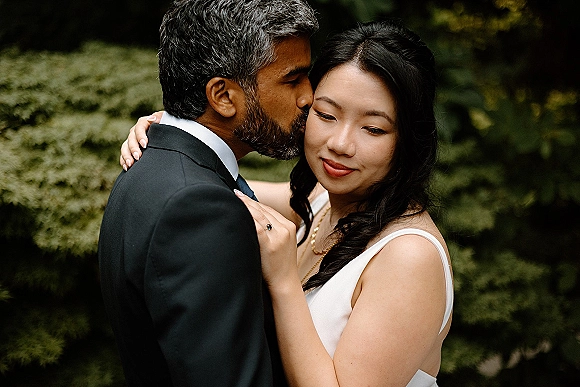 Couple portrait with groom kissing bride’s forehead as they embrace, her engagement ring visible against lush garden foliage backdrop