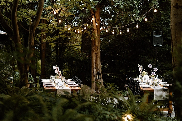 Outdoor reception tablescape with wooden farmhouse reception tables, white runner, floral centerpieces, lantern, and string lights in a forest setting