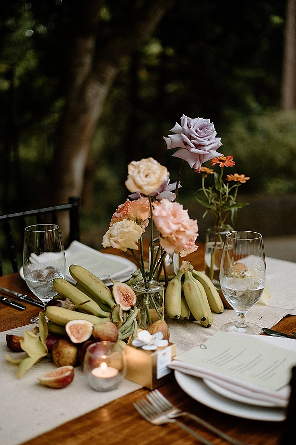 Reception tablescape on a wooden outdoor reception table with bud vases of roses and carnations, fruit centerpiece with bananas and figs, and votive candles