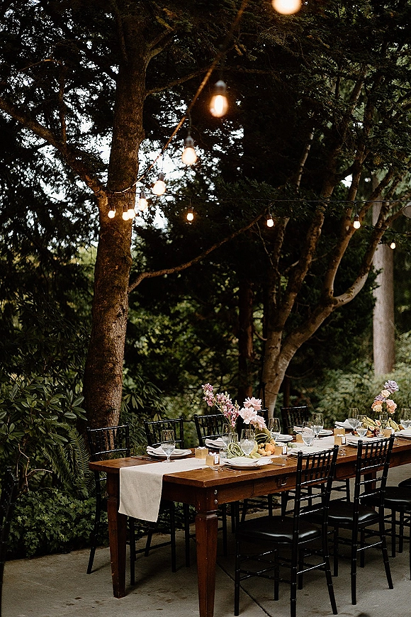 Outdoor reception tablescape with a wood farmhouse table, black chairs, pastel floral centerpieces, and string lights on a garden patio
