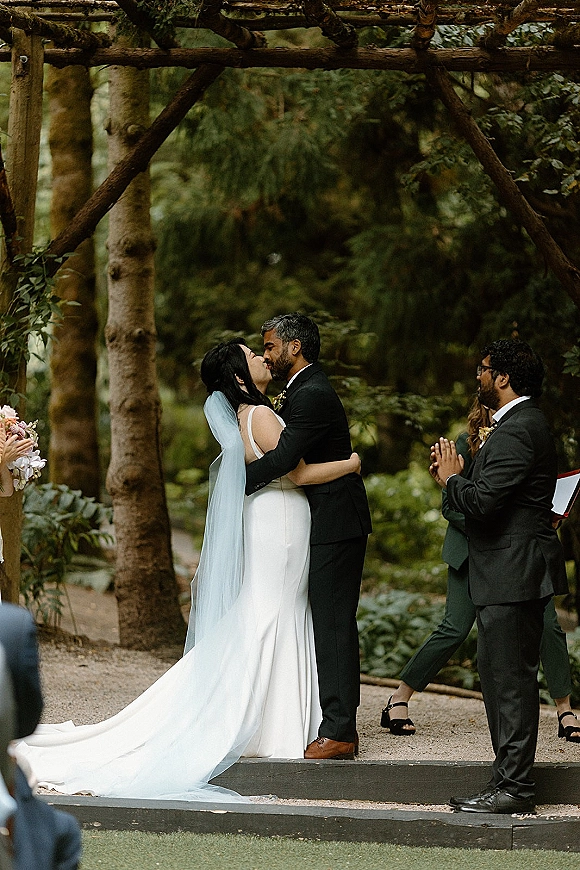 Ceremony kiss at an outdoor wedding ceremony as bride in veil and groom in black suit kiss under a wooden arbor in the forest greenery