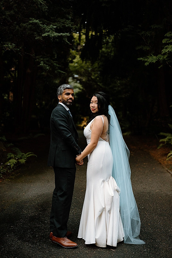 Couple portrait of bride and groom holding hands on a wet forest road, bride in satin dress and long veil looking back in moody light