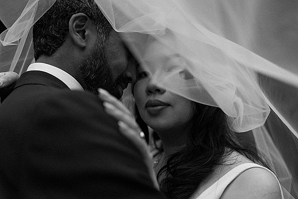 Wedding couple portrait of bride and groom under veil, touching foreheads outdoors, her hand on his face showing a wedding ring