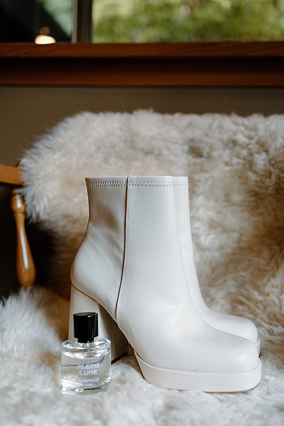 Bridal shoes, white wedding boots with block heels beside a perfume bottle on a faux fur chair by a window with greenery outside
