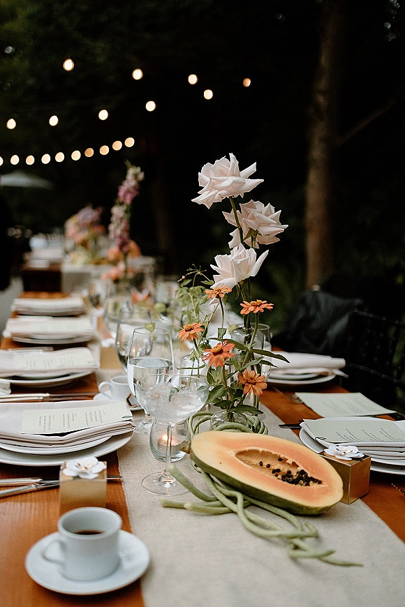 Reception tablescape on a long wooden outdoor reception table with linen runner, bud vases of white roses and orange flowers, tea lights, and string lights in a garden at dusk