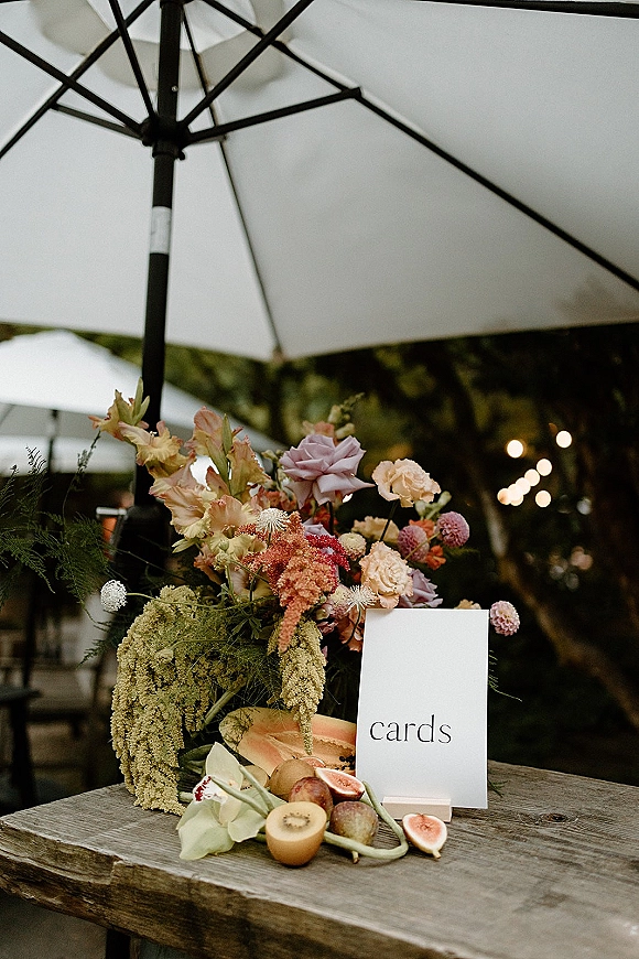 Wedding cards table with a wedding cards sign beside pastel florals and fruit accents on a rustic patio under string lights and umbrellas