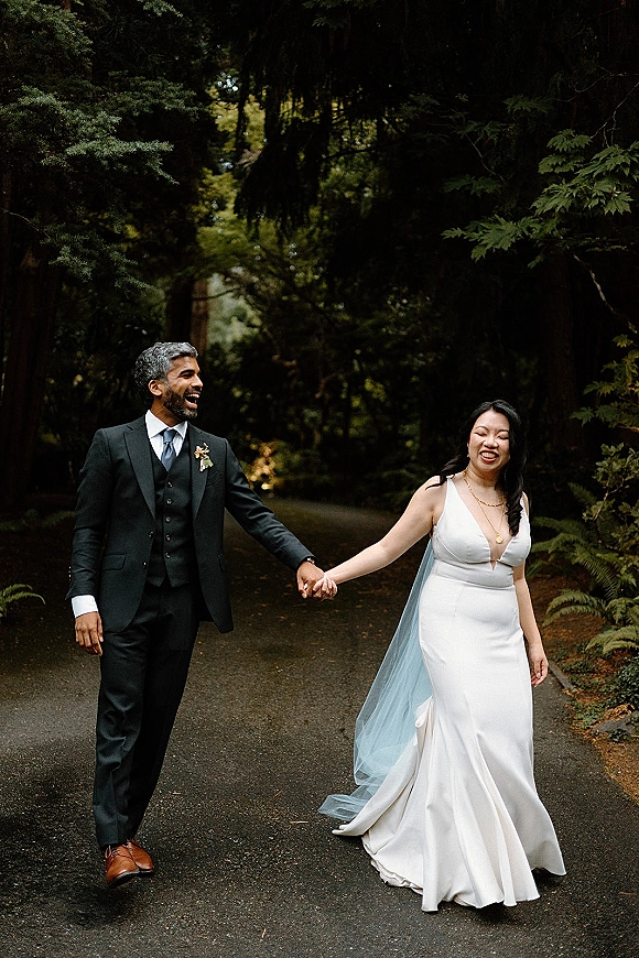 Couple portrait of bride and groom holding hands, walking on a paved forest road, her long veil trailing behind amid ferns