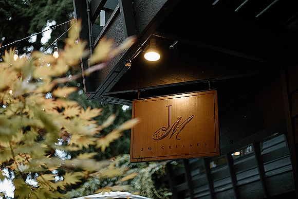 Venue sign hanging with logo lettering under warm string lights on a rustic building exterior, framed by leafy trees at dusk