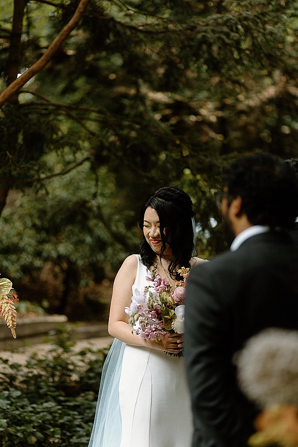 First look moment as groom in black suit sees bride in sleeveless gown holding a colorful bouquet on a garden walkway, light blue veil accent
