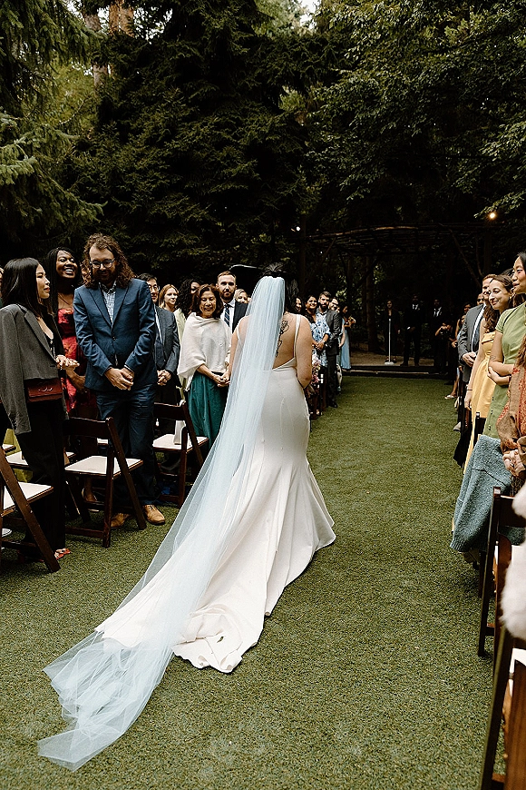 Processional moment as the bride walking down aisle, seen from behind with cathedral veil train, guests beside wood folding chairs in a garden setting