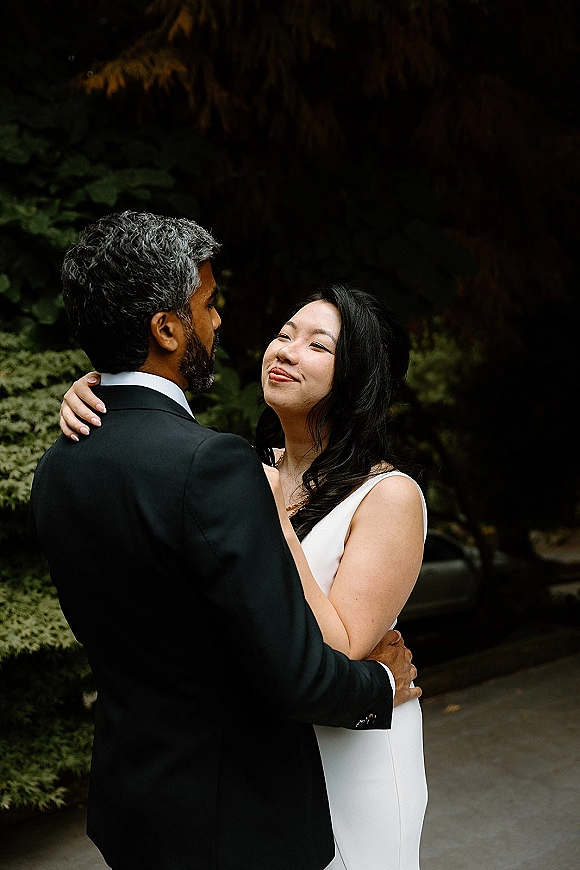 Couple portrait of bride and groom hug on an outdoor path, her sleeveless white dress and his black suit framed by garden trees