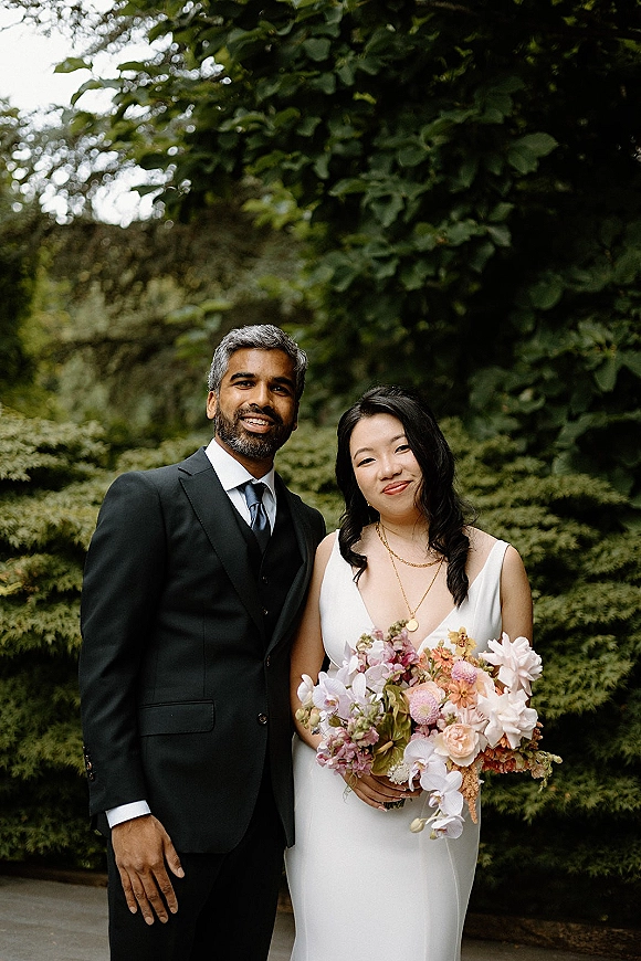 Couple portrait of bride and groom portrait posing on a garden pathway, bride in simple slip gown holding a colorful bouquet amid greenery